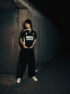 A person stands in a dark alleyway wearing a black Real Madrid soccer jersey with white sponsor logos, black pants, white sneakers, and a black beanie.