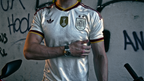 A man wearing a white Spain national football team jersey with gold and maroon trim, standing against a graffiti-covered wall.