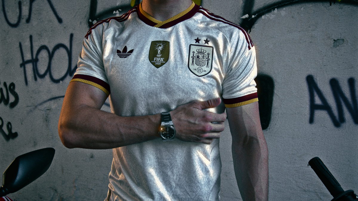 A man wearing a white Spain national football team jersey with gold and maroon trim, standing against a graffiti-covered wall.