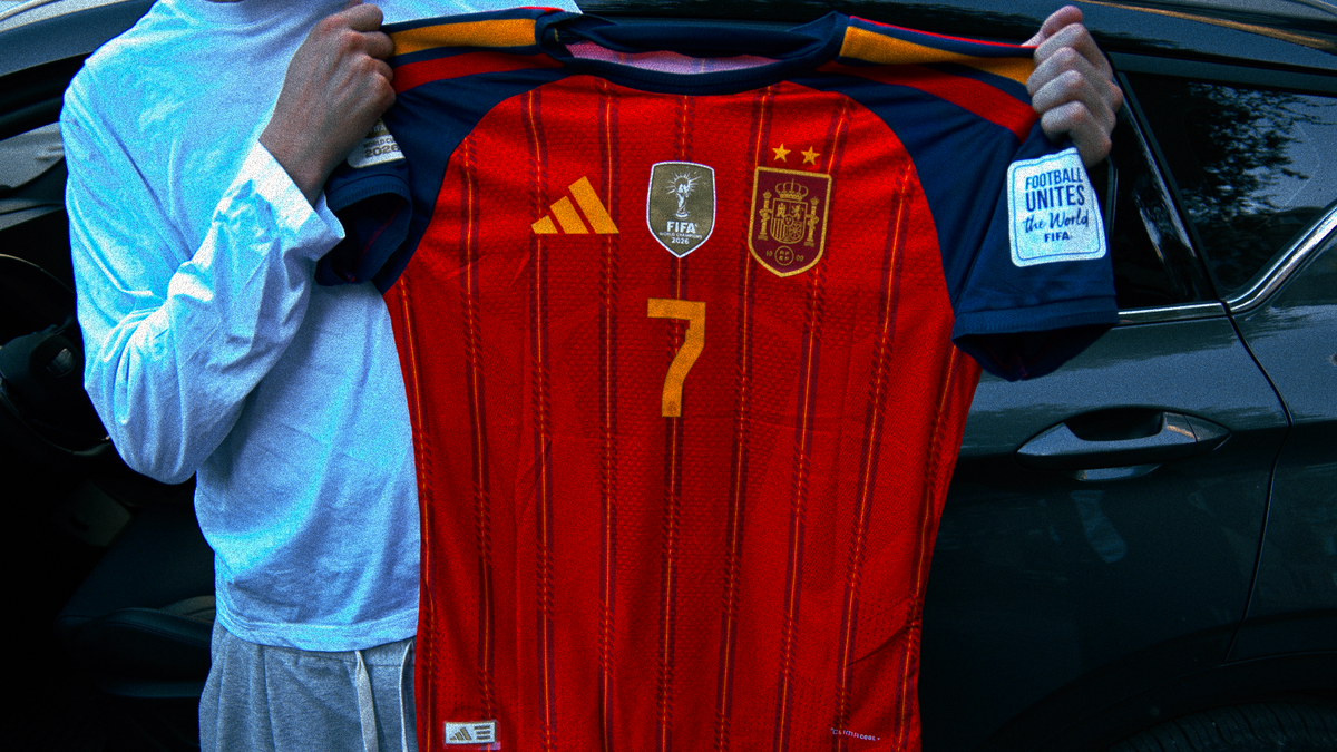 A person holds up a red Spain national football team jersey with the number 7, featuring navy sleeves, in front of a dark car.