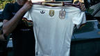 A person holds up a white Spain national team soccer jersey with gold and maroon trim, featuring the logo, FIFA patch. The photo is taken outdoors in front of a dark vehicle.