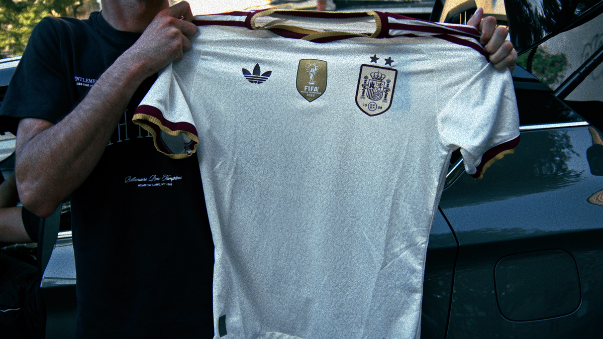 A person holds up a white Spain national team soccer jersey with gold and maroon trim, featuring the logo, FIFA patch. The photo is taken outdoors in front of a dark vehicle.